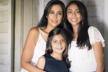 Outdoor portrait of an attractive mother and her daughters. The smiling mother and the older sister in white top, the cute little girl in black top standing in front of the door.
