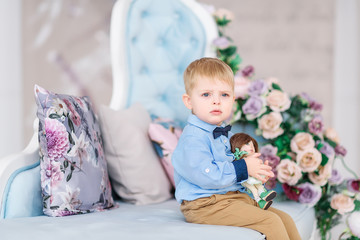 Fototapeta premium Portrait of a small cute boy gentleman in a blue shirt and with a tie. The child stands near a large window in a bright cozy house and holds a bouquet of flowers in his hand - a gift for the mother