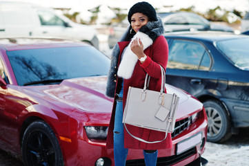 Rich african american girl in red coat and fur against red muscle car. Black stylish successful businesswoman.