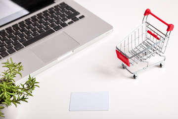 Online shopping. Bank card nearby a laptop and mini shopping cart on white background top view.