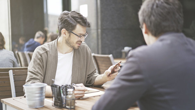 A man in eyeglasses holding looking at the phone at cafe outdoors. A man dressed in a jacket wearing eyeglasses listening to his friend talking with a phone in hand