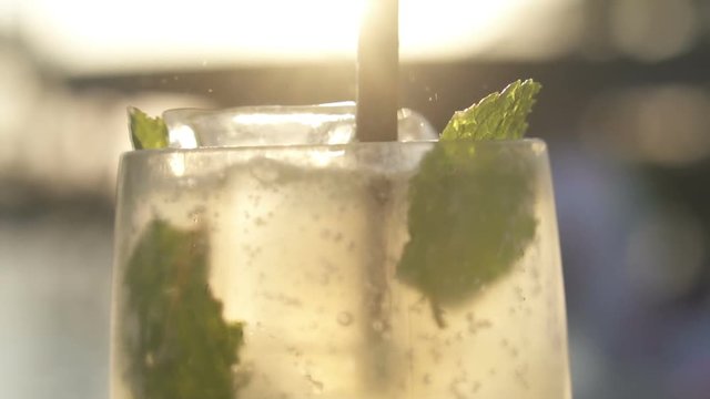 Closeup glass of Mojito cocktail with mint leaves and straw over beautiful sunset background. Sparkles and bubles video in slow motion