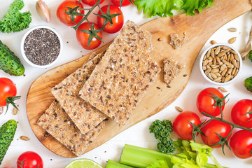 Top view of whole grain rye crispbreads on a wooden serving board surrounded fresh vegetables, greens and seeds over white background.