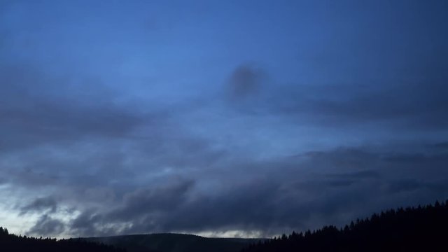 4K Time-Lapse. Nubes bajas y altas pasando r&aacute;pidamente de izquierda a derecha a lo largo de una monta&ntilde;a con bosque de pinos al caer la noche. 