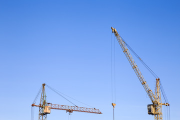Fototapeta na wymiar Front distant view of two cranes on a light blue sky background. One crane`s jib-arm is horizontal, the other one`s is placed diagonally with the lower sheave and hook hanging