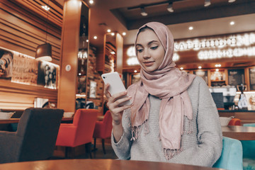 cute beautiful Arab girl in hijab sits in cafe, looks into her smartphone, oriental style