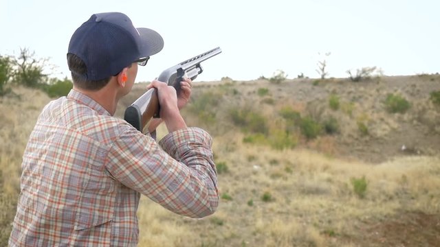 Man shooting a shotgun in the desert in 4k. The tall white male follows the clay pigeon with the barrel of his gun, pulls the trigger and expels the used shell from the pump action firearm.