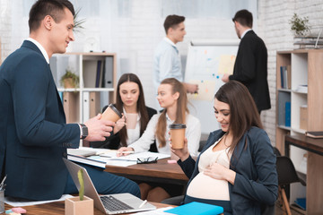 Pregnant girl in office is drinking coffee with chief in office. Pregnant with colleagues.