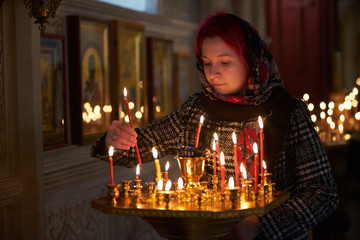 Praying young woman with candle near pedestal with many other candles in church by her hand to show her faith and esteem to God