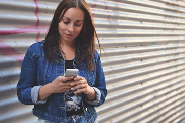 A hipster girl dressed in urban style stands against the wall with a phone in her hands. The teenager dials a text message in the messenger. Life style. Communication. Modern tendencies.