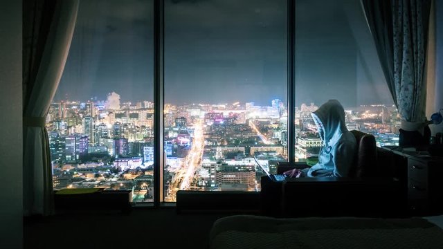 Hooded anonymous man working on laptop computer in dark hotel room, panoramic window with illuminated night city view in background. Timelapse, 4K UHD. Zoom in.