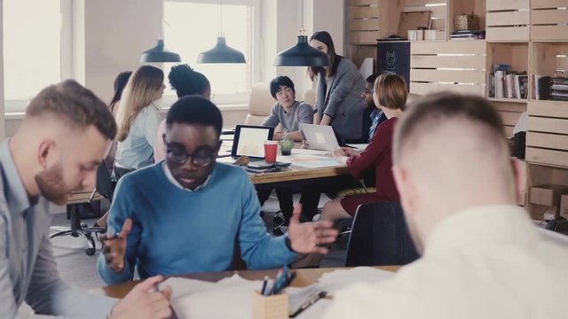 Female team leader gives new tasks to employees. Happy multiethnic colleagues sit by the table at office meeting 4K.