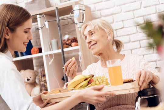 Girl Is Caring For Elderly Woman At Home. Girl Brings Breakfast On Tray. Woman Is Eating Fruit.