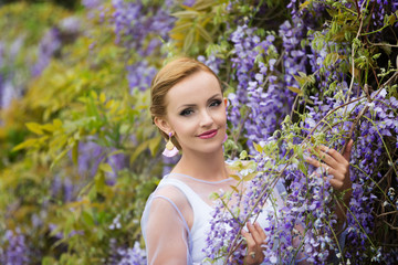 A portrait of young Caucasian woman with blond hair near purple wisteria, looking straight to camera.