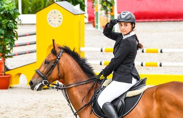 Beautiful brunette girl in uniform and bay dressage horse at showjumping competition. The salute to the judge. Equestrian sport background.