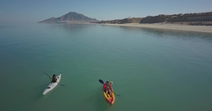 Aerial of two women kayaking toward the camera in Baja with a mountain in the distance. One woman in bikini with a dog.