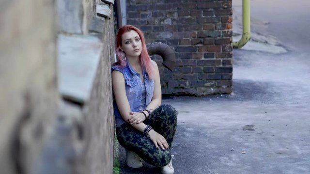 Teenage girl with nose and lips piercing and pink hair sit leaning against brick wall and looking at camera. Slow motion. HD