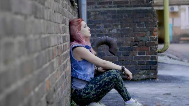 Teenage girl with nose and lips piercing and pink hair sit leaning against brick wall. Slow motion. HD
