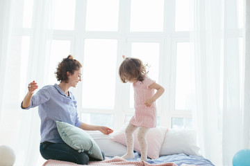 Fototapeta premium Young mother and her little daughter jumping on bed. Funny pillow fight. Soft pastel colors, pink and blue. Selective focus. Play together and enjoy the moment! Family time on weekend. 