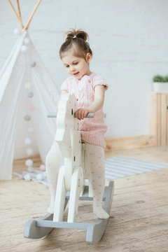 Little girl in pink dress riding wooden toy horse and smiling. Cute daughter with present. Nursery room interior with wooden horse and tent lodge. Soft pastel colors. 
