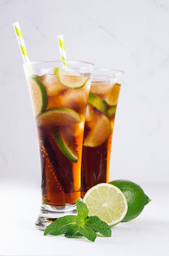 Summer fresh carbonated cola with ice in two long glasses with mint and lime on white wood board, closeup, vertical.
