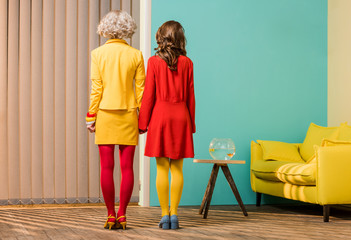 back view of women in bright retro styled clothing holding hands at colorful apartment, doll house...