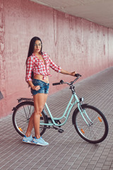 Fototapeta na wymiar Sexy stylish brunette girl wearing a pink flannel shirt and denim shorts in glasses, posing with city bike against a pink wall.