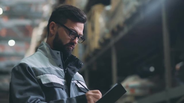 Storekeeper in the warehouse make an audit and an inventory. Man at a large storage, looks around, finds a box and takes notes. Modern man with a beard is dressed in gray uniform, works in a warehouse
