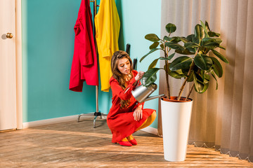 beautiful woman in red dress squatting and watering plant with watering can at home