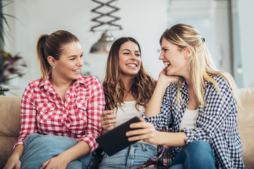 Fototapeta premium Three best friends using digital tablet together. Women sitting on sofa having fun surfing on the internet using smart digital pc tablet modern device