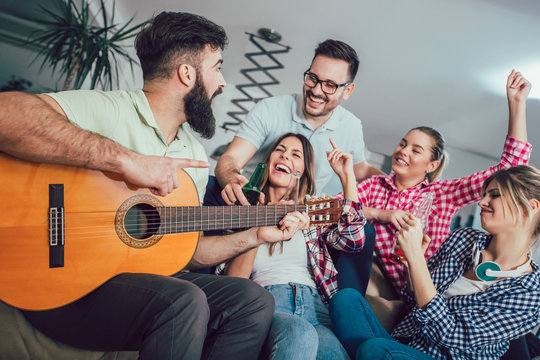 Group of happy young friends with guitar having fun and drinking beer in home interior