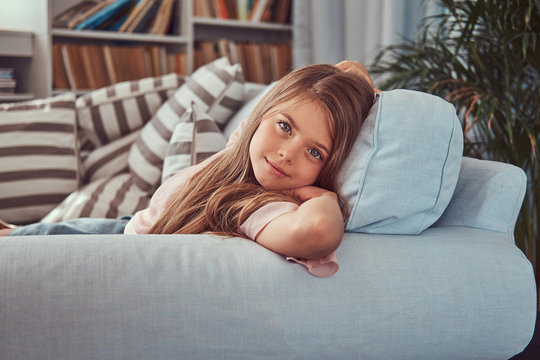 Portrait of a smiling little girl with long brown hair and piercing glance, lying on a sofa at home