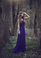 Fototapeta premium Girl elf with long hair and blue eyes in tiara and a long blue dress with a train walking through the spring blossoming forest. Girl Princess dreams.