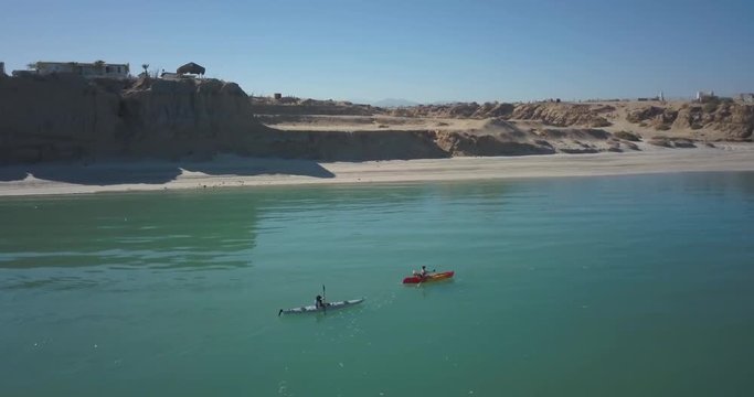 Aerial camera backing away as two kayaking women paddle along a sandy shore with cliffs over calm water.
