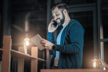 Business talk. Low angle of optimistic vigorous IT guy rising tablet while speaking on phone and laughing