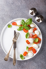 White plate of healthy classic delicious caprese salad with ripe tomatoes and mozzarella cheese with fresh basil leaves on gray concrete background with space for text. Italian food. Top view.