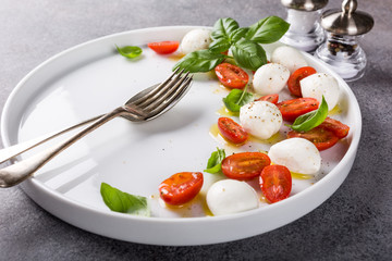 White plate of healthy classic delicious caprese salad with ripe tomatoes and mozzarella cheese with fresh basil leaves on gray concrete background with space for text. Italian food.