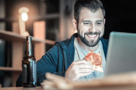 Time for break. Joyful optimistic IT guy chewing pizza slice while looking down and grinning