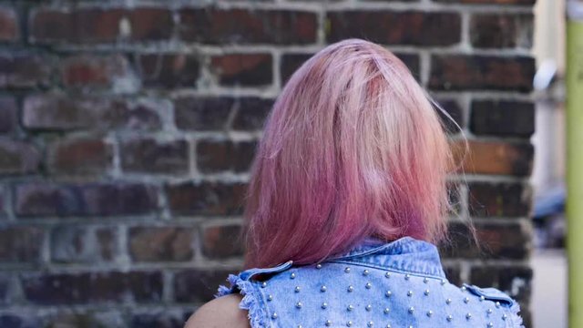 View from the back of young trendy teenage girl with pink long hair walking along brick house. Slow motion. HD