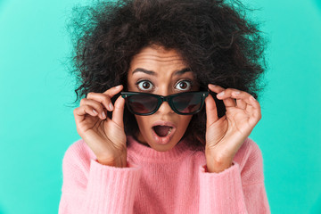 Photo of surprised woman 20s with shaggy hair looking on camera with excitement from under black sunglasses, isolated over blue background