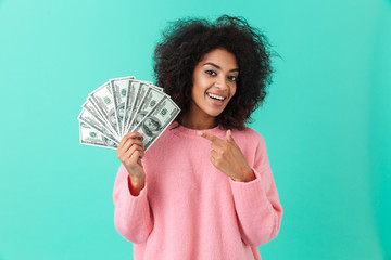 Portrait of excited woman 20s with afro hairstyle pointing finger on fan of money dollar bills,...