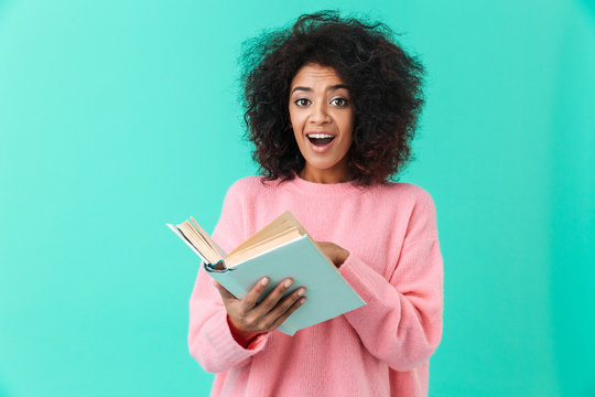 Image of happy woman 20s with afro hairstyle looking on camera with excitement and holding book in hands, isolated over blue background