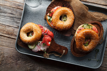 Bagels with salmon fish, cream cheese, cucumber and fresh radish slices on metallic tray on rustic gray wooden background