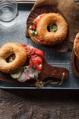 Bagels with salmon fish, cream cheese, cucumber and fresh radish slices on metallic tray on rustic gray wooden background