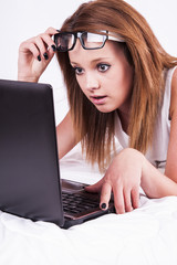 Natural looking young woman with glasses lying down in bed, searching internet on notebook and being surprised