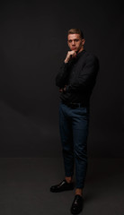 attractive white man in black shirt and blue pants stands on a dark background