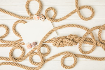 top view of empty paper with seashells on brown nautical knotted ropes on white wooden surface