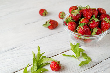 Glass plate with ripe red strawberries and green mint leaves on a white wooden table. Strawberries scattered on a table.