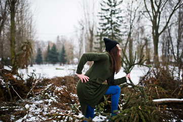Young girl wear on long green sweatshirt, jeans and black headwear at  branches of the pine tree in winter day.