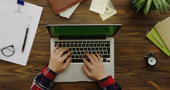Top view on the female hands with the motley sleeves working on the laptop computer with a green screen on the wooden table with office stuff on it. Chroma key.